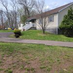 Small, light-colored house with a lawn and surrounding trees on a bright day.