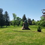 Bronze statue on a pedestal in a sunny, grassy park with trees.