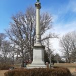 Statue on tall column in a park setting with bare trees under a blue sky.