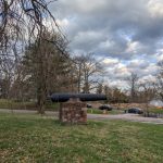 Old cannon mounted on a brick pedestal in a grassy park setting under a cloudy sky.