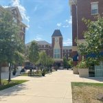 Campus view with brick buildings, central clock tower, and trees lining a walkway under blue sky.