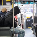 Person in black jacket stands inside a public transit bus, looking out the window.