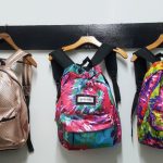 Three colorful backpacks hanging on wooden hangers against a white wall.