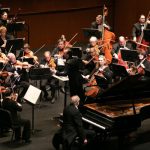 Orchestra performing on stage with a grand piano in the foreground.