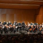Orchestra performing on stage before a large audience in a concert hall.