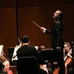 Conductor leads orchestra performance on stage under dramatic lighting.