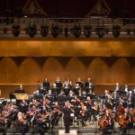 Orchestra performing on a large stage with dramatic warm lighting.