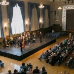 Orchestra performs on a stage in a large, ornate concert hall before an audience.