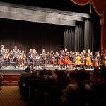 Orchestra performs on a stage before an audience in a grand hall.