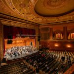 Ornate theater interior with a large audience seated for an event under a domed ceiling.