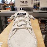 White Sperry low-top sneakers displayed on a wooden counter with pricing signage in the background.