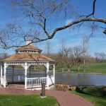 Victorian gazebo by a pond with trees and green parkland under blue sky.