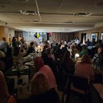 Crowded indoor event with people seated at tables, suggesting a dinner or gathering.