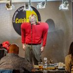 Comedian in red sweater performs on stage at a comedy club.