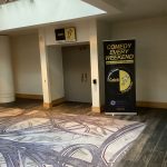 Modern hotel lobby with a large patterned rug, high ceilings, and promotional signage.