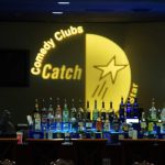 Comedy Clubs Catch logo displayed above a bar stocked with various liquor bottles.