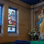 Stained glass windows illuminate a church interior with a statue of the Virgin Mary.