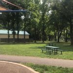 Park scene with grassy area, picnic tables, and a small building under tall trees.