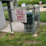 Warning sign posted near a chain-link fence next to a concrete structure and sports field.