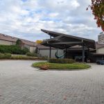 Modern building entrance with covered walkway, landscaping, and paved driveway under a cloudy sky.