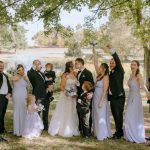 Joyful wedding party celebrating outdoors under green trees.