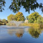 Fountain spraying water in a calm pond, surrounded by autumn trees under a blue sky.