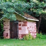 Small, rustic stone and stucco shed nestled under leafy green trees in a grassy area.