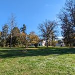 Autumn trees frame a white farmhouse under a bright blue sky on a grassy field.