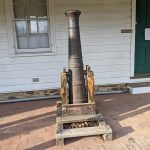 Old, rusted metal chimney pipe mounted on a wooden stand outdoors near a white building.