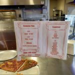 Menu board displaying tomato pies and toppings prices at a food service counter.