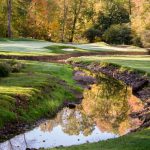 Golf course green with a winding stream reflecting autumn trees.