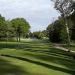 Green golf course with sprinkler system spraying water under a partly cloudy sky.