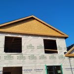 New house under construction with exposed wood framing and white sheathing against a clear blue sky.