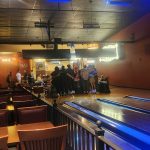 Crowded bar interior with a long, illuminated wooden counter and patrons socializing.