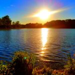Sunset over calm lake with bright sun reflection on the water and dark trees lining the shore.