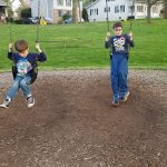 Two boys swing on playground swings in a grassy park area.
