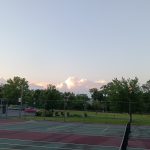 Outdoor tennis court under a pale sky with scattered clouds at sunset.