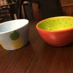 Two colorful bowls, one with a pineapple design and another bright orange/green, on a dark wooden table.