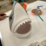 Sculpting a large, white shark model on a white table with art supplies nearby.