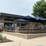 Outdoor dining area with dark umbrellas in front of a modern building under a bright sky.