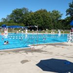 Sunny outdoor public pool with people swimming and sunbathing under blue skies.