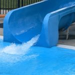 Blue water slide empties into a bright blue swimming pool with splashing water.