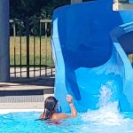 Child slides down blue water slide into a pool on a sunny day.