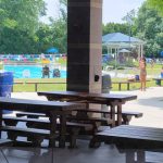 Outdoor patio area overlooking a sunny public swimming pool and park.
