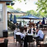 Outdoor dining area by the water with tables, chairs, and umbrellas on a sunny day.