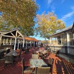 Outdoor patio dining area with wooden deck, tables, and trees under a blue sky.