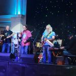 Musicians perform on a stage with blue lighting and starry backdrop.