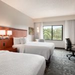 Modern hotel room with two beds, desk area, and large window overlooking trees.