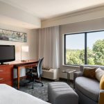 Modern hotel room with a sofa, desk area, and large window overlooking green trees.
