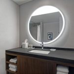 Modern bathroom vanity with dark countertop, round backlit mirror, and wood storage.